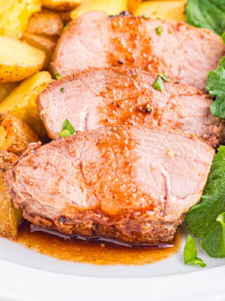 Three slices of cooked pork tenderloin with balsamic sauce, served with roasted potatoes and a side of leafy greens on a plate.