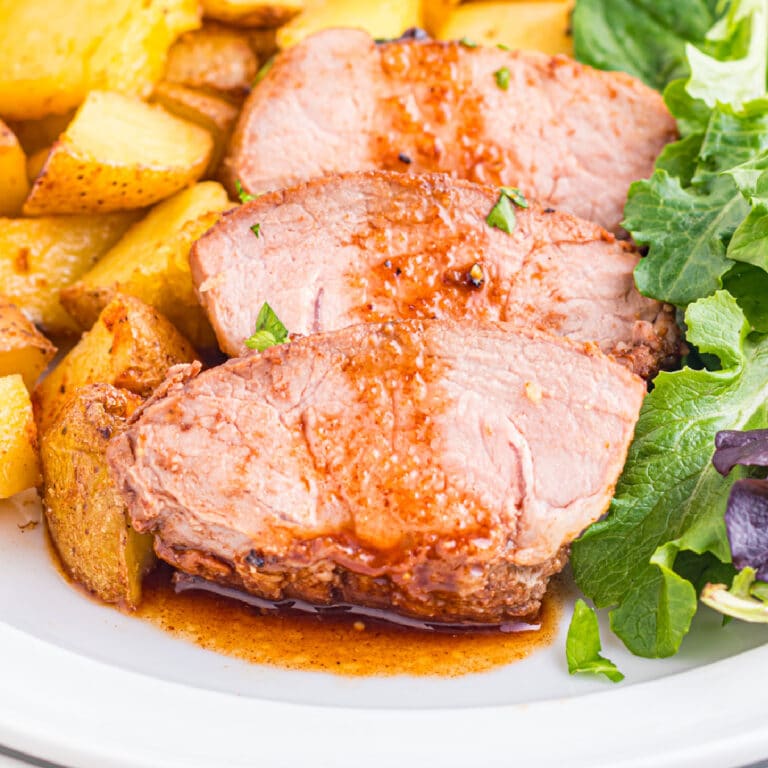 Three slices of cooked pork tenderloin with balsamic sauce, served with roasted potatoes and a side of leafy greens on a plate.