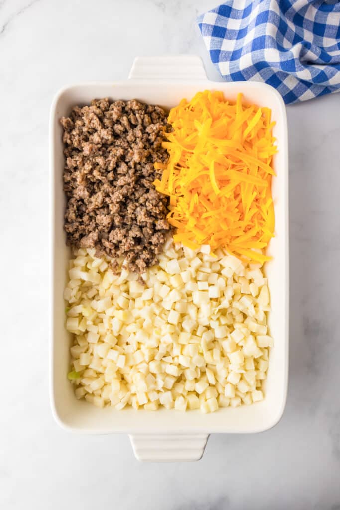 A baking dish with diced potatoes, cooked ground sausage, and shredded cheddar cheese portioned in a casserole dish before mixing.