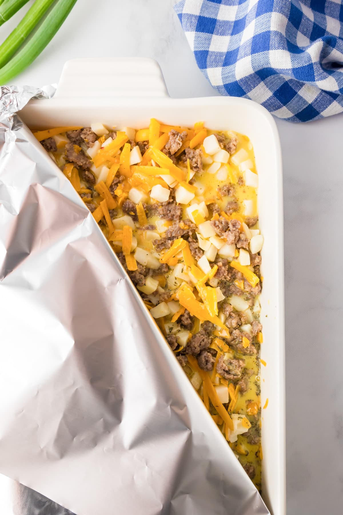 A baking dish partially covered with aluminum foil full of a uncooked sausage potato breakfast casserole.