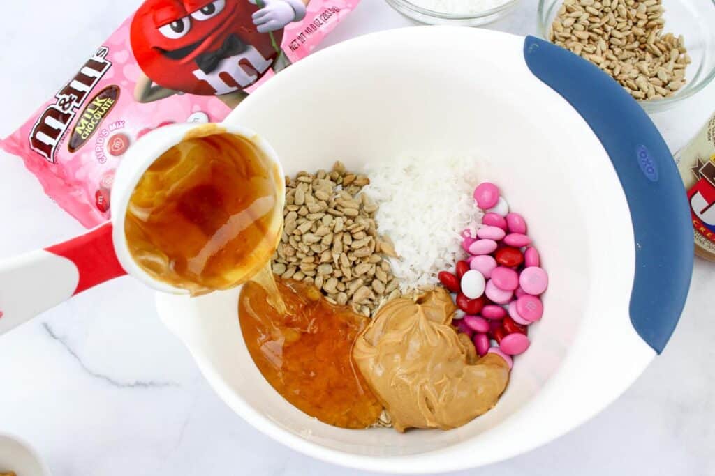 Bowl containing peanut butter, coconut flakes, sunflower seeds, and pink and red candies with honey being poured from a measuring cup into the bowl.