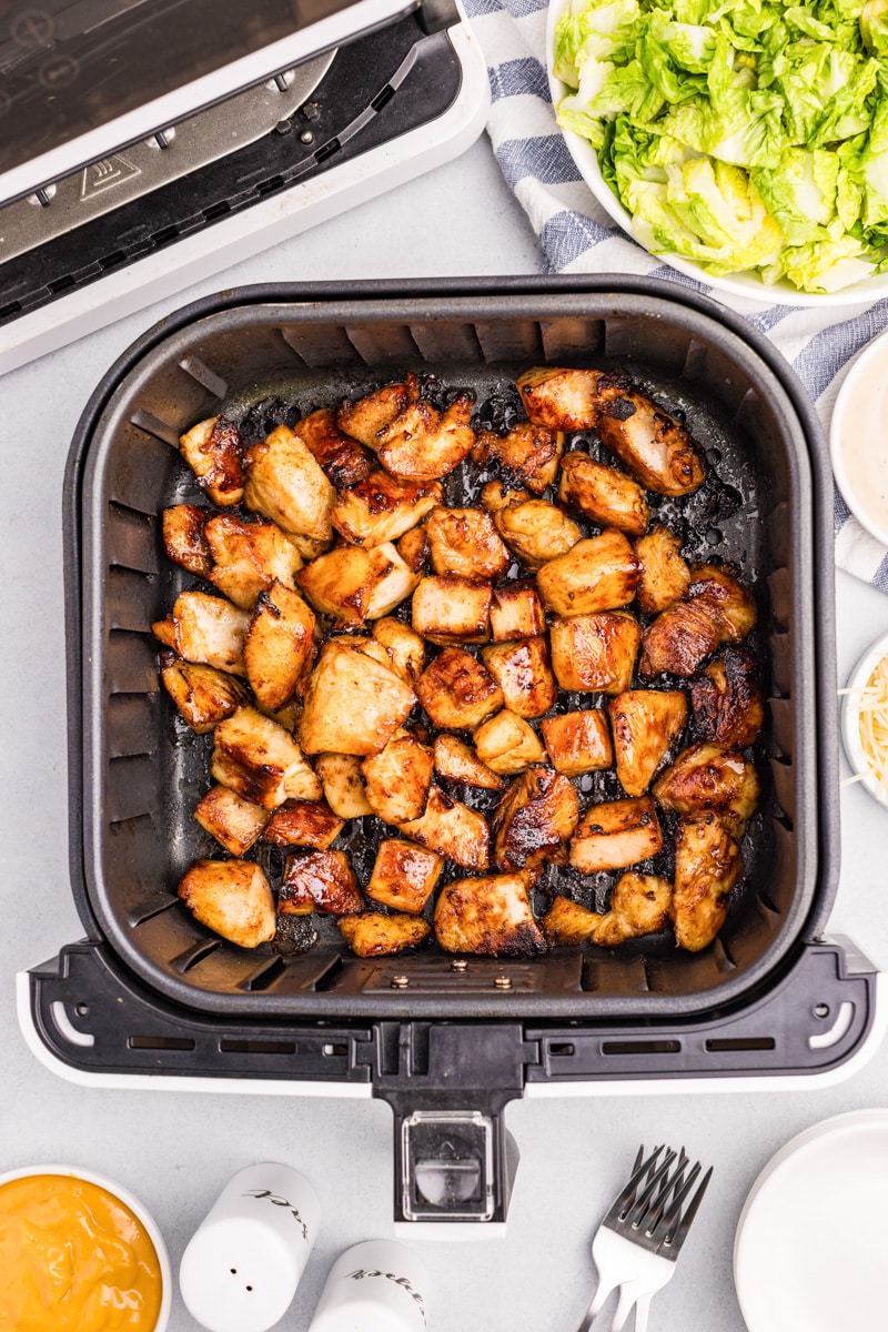 Cooked golden brown air fryer chicken chicken bites in an air fryer basket with salad, plates and other dinner items nearby.