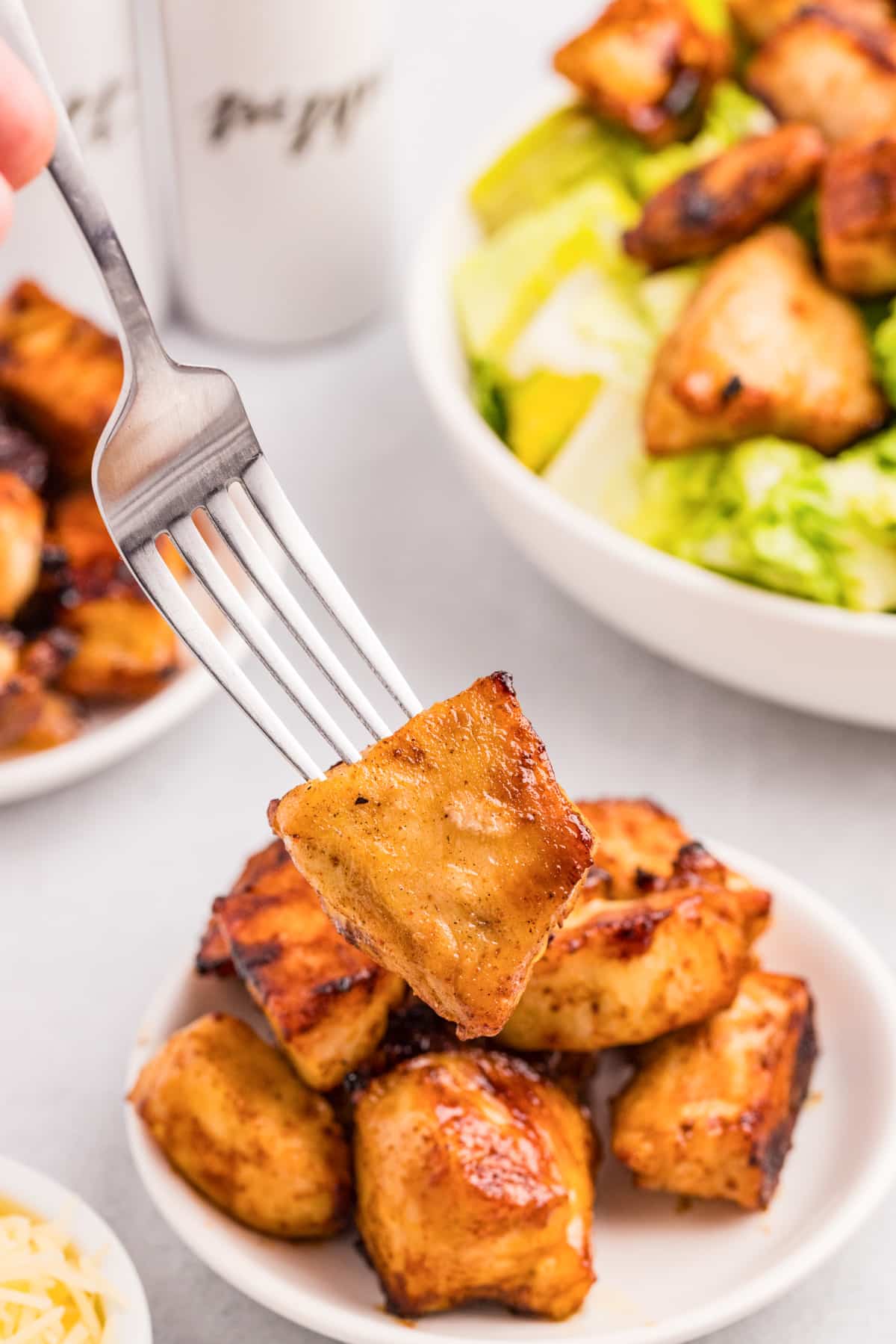 A fork holding a piece of air fryer chicken bites pulled from a plate full with more on a salad in the background.