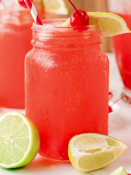 A mason jar filled with cherry limeade surrounded with pieces of lime and a cherry on top.