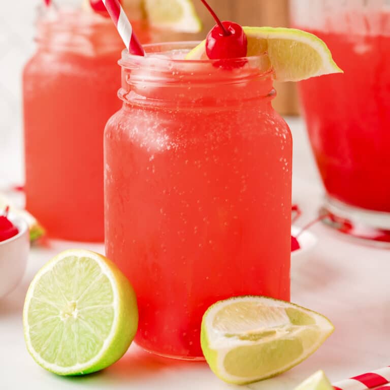 A mason jar filled with cherry limeade surrounded with pieces of lime and a cherry on top.