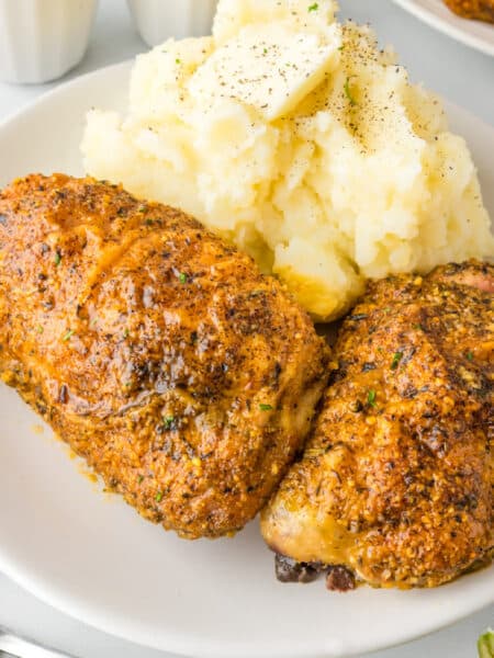 Square view of a plate of two roasted garlic parmesan chicken thighs next to a serving of mashed potatoes with butter.