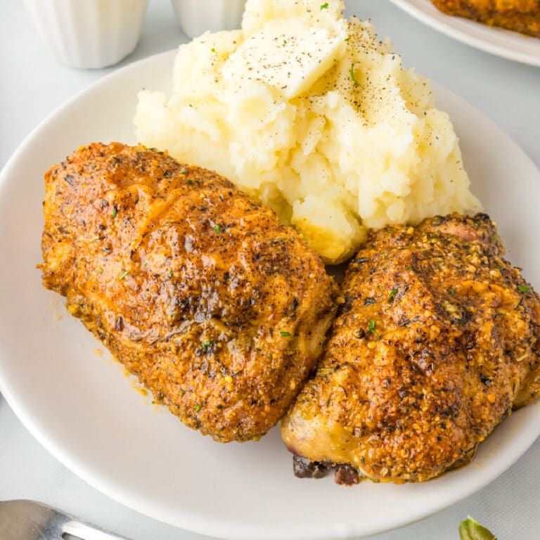 Square view of a plate of two roasted garlic parmesan chicken thighs next to a serving of mashed potatoes with butter.