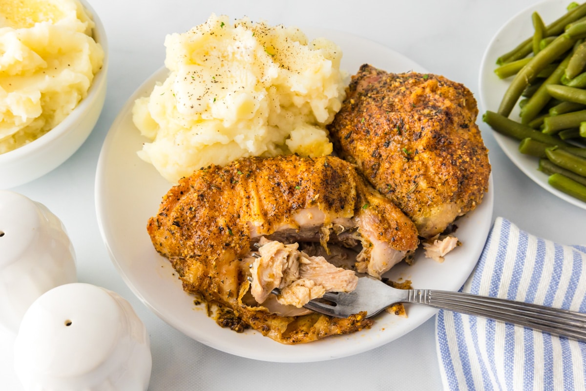 Wide view of a plate with two pieces of roasted garlic parmesan chicken thighs with potatoes and green bean sides and a fork pulling a bite from the chicken.