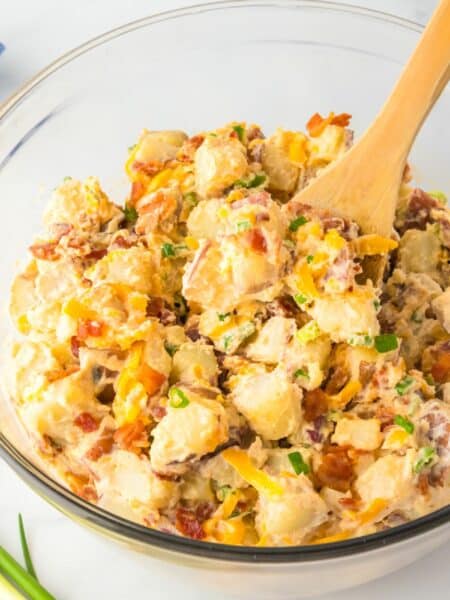 Large bowl of potato salad with bacon, cheese and green onion in a creamy dressing with a wooden spoon stirring.