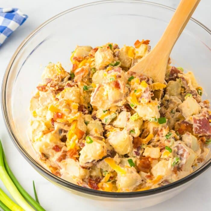 Large bowl of potato salad with bacon, cheese and green onion in a creamy dressing with a wooden spoon stirring.