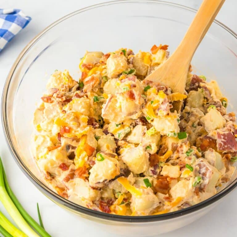 Large bowl of potato salad with bacon, cheese and green onion in a creamy dressing with a wooden spoon stirring.