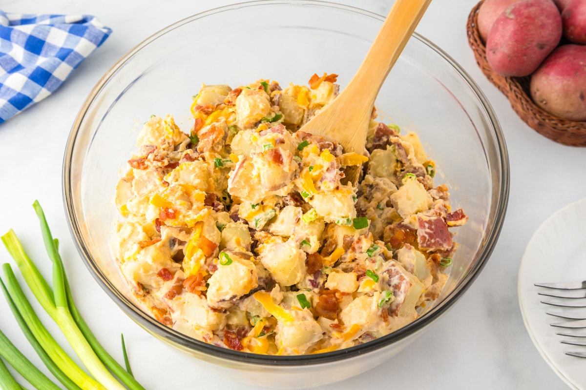 Wide view of a glass bowl of loaded baked potato salad with bacon pieces, green onion and cheese, mixed with a wooden spoon.