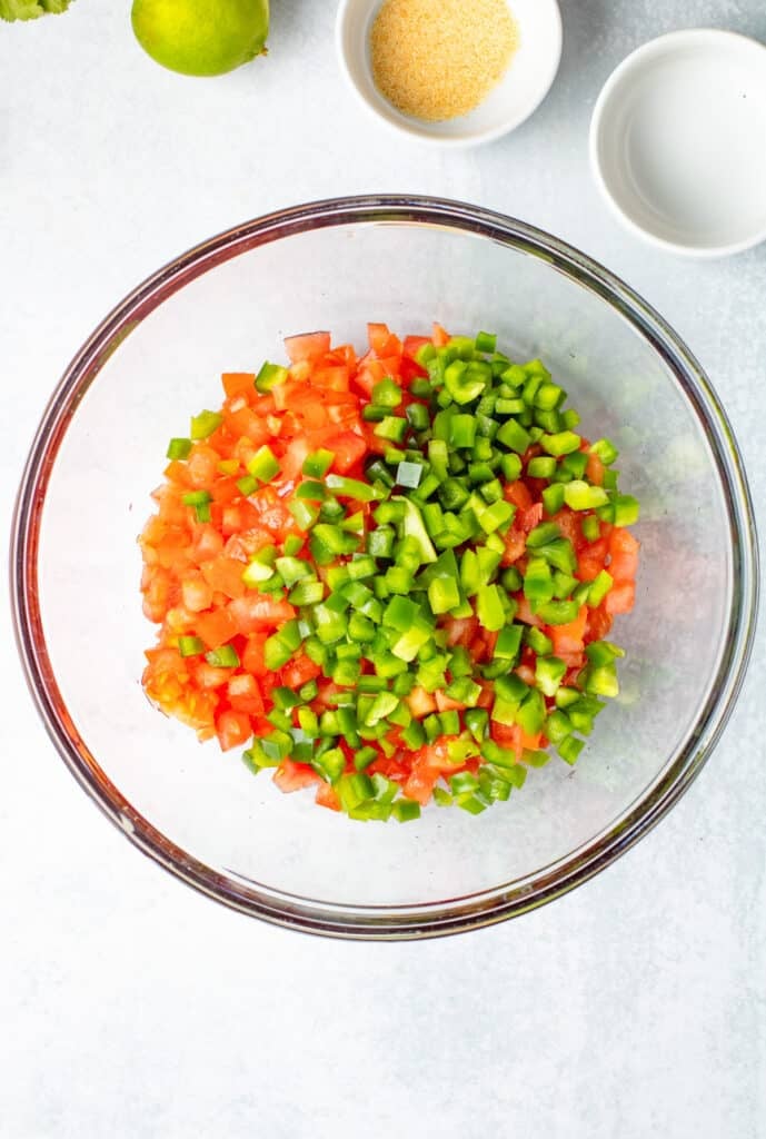 Chopped tomatoes and green bell peppers in a glass bowl, with small bowls of seasoning for making Pico de Gallo.