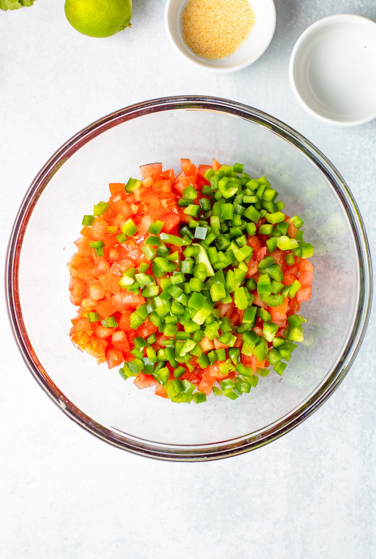 Chopped tomatoes and green bell peppers in a glass bowl, with small bowls of seasoning for making Pico de Gallo.