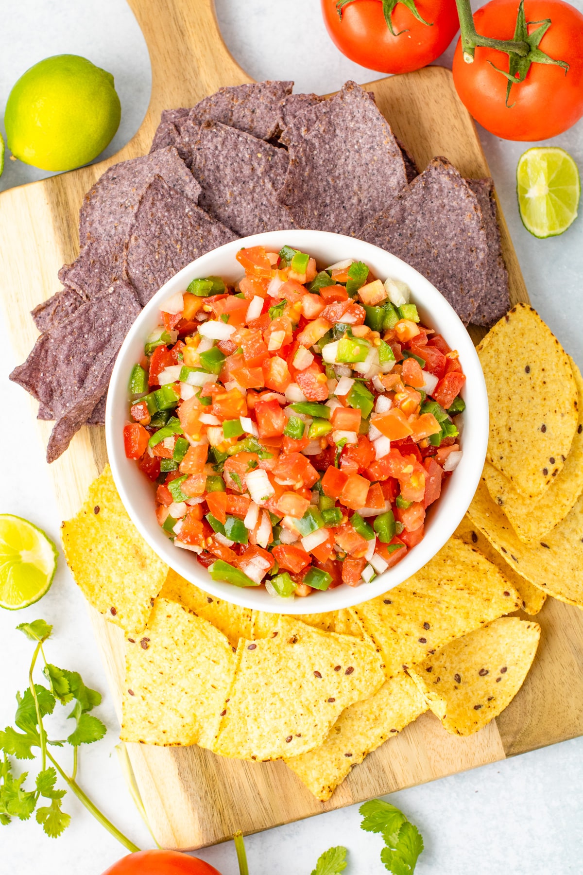 A bowl of Pico de Galo surrounded by blue and yellow corn tortilla chips on a wooden board from above.