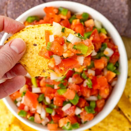 A hand holding a tortilla chip after dipping in the bowl of Pico de Galo below.