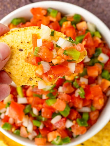 A hand holding a tortilla chip after dipping in the bowl of Pico de Galo below.