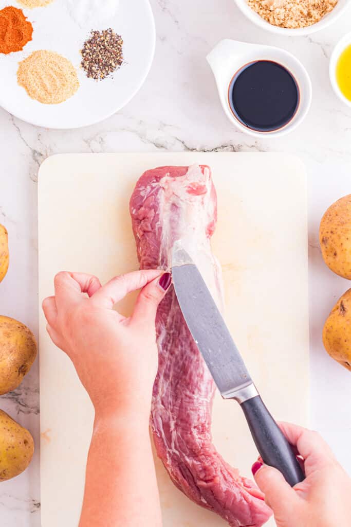 A person's hands trimming the silver skin off pork tenderloin with a knife and other ingredients for pork tenderloin and potatoes near the cutting board.