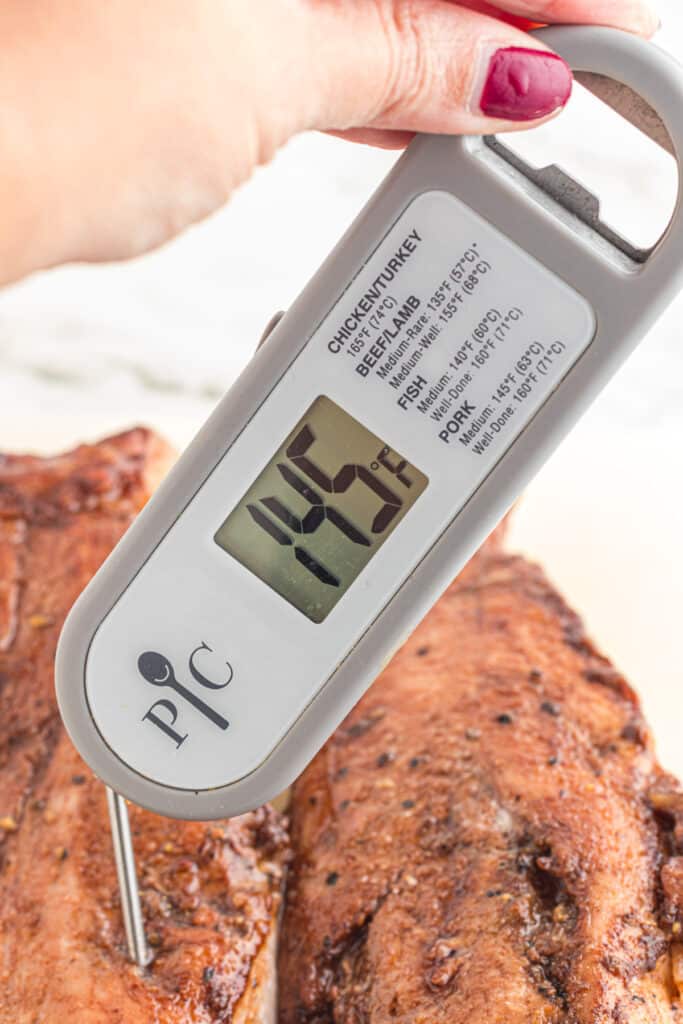 A hand holding a meat thermometer inserted into pork tenderloin showing it is 145 degrees.