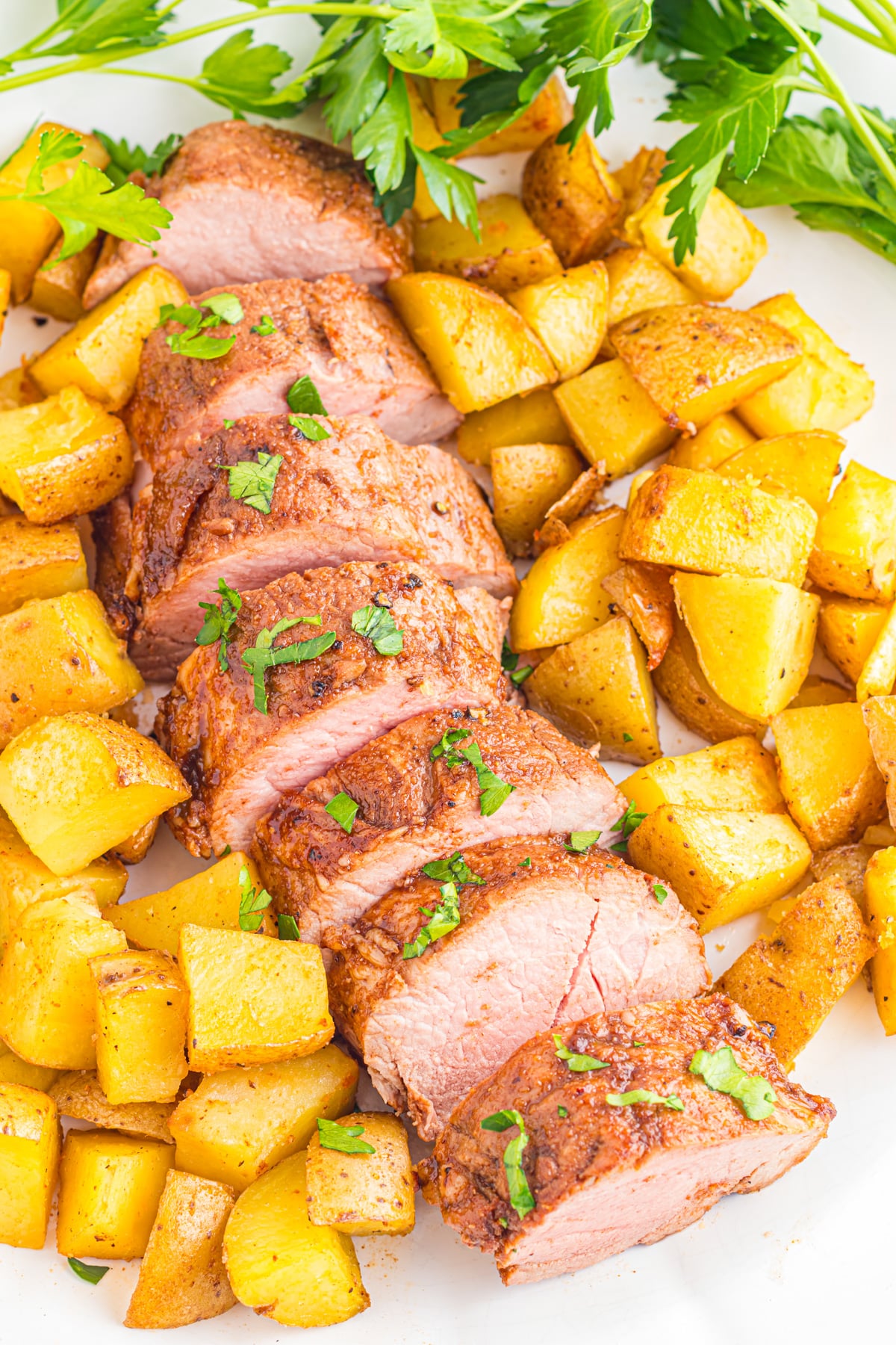 Sliced roasted pork tenderloin garnished with herbs, surrounded by roasted potato cubes on a platter.