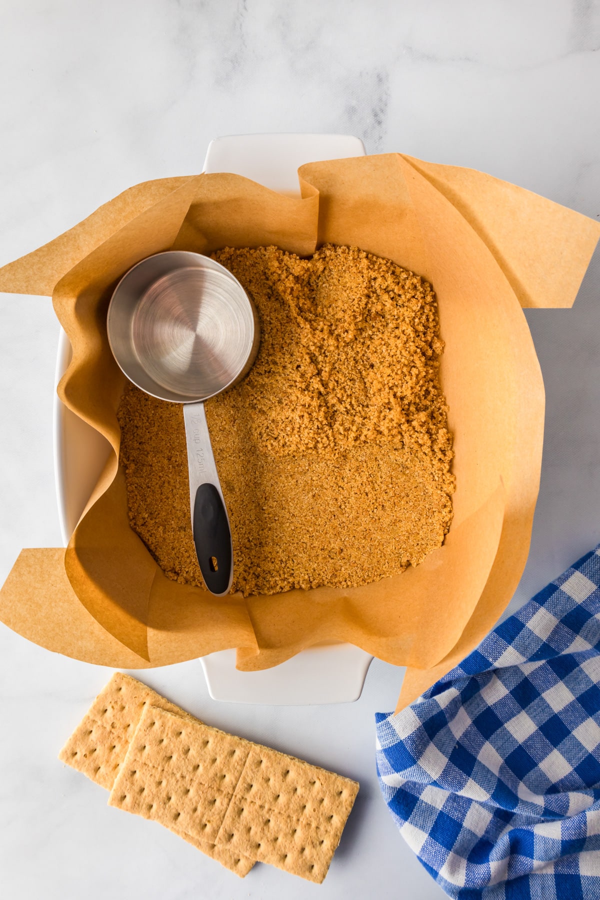 Baking pan lined with parchment paper with a graham cracker crust being pressed into the bottom of the pan with a measuring cup for strawberry cheesecake bars.