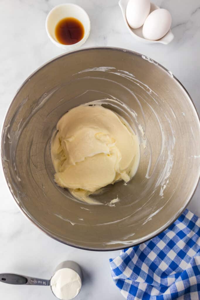 A metal bowl with creamed cream cheese and sugar with eggs , vanilla and sour cream nearby for strawberry cheesecake bars.