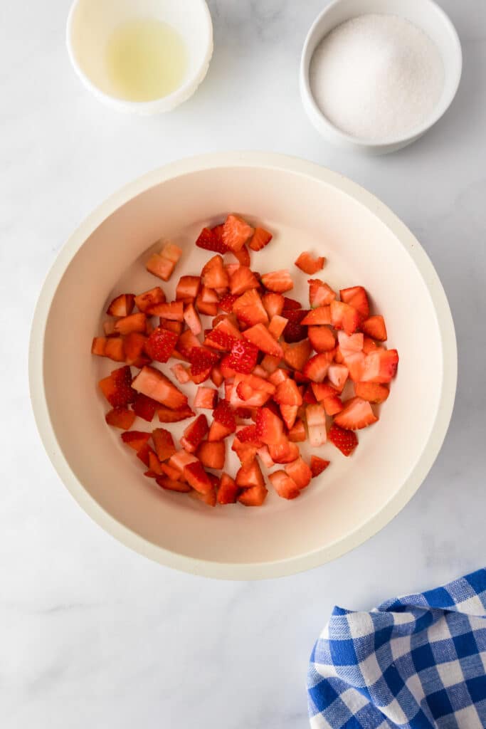 Chopped strawberries in a bowl, a cup of sugar, and a bowl of liquid on a marble surface with a blue checkered cloth.