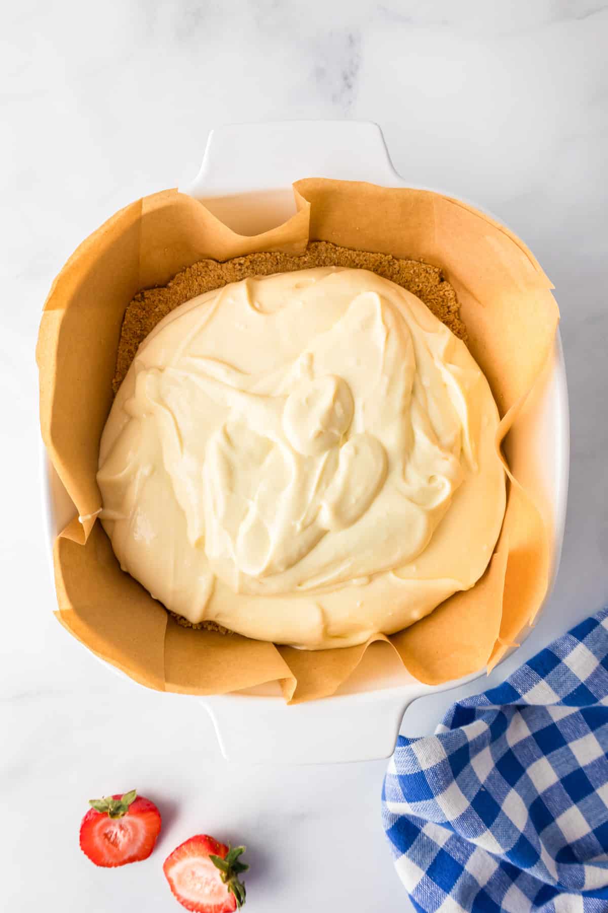 Cheesecake batter spread over a graham cracker crust in a parchment-lined square dish from above.