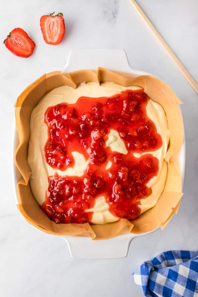 A baking dish lined with parchment paper contains cheesecake batter topped with strawberry sauce before mixing.