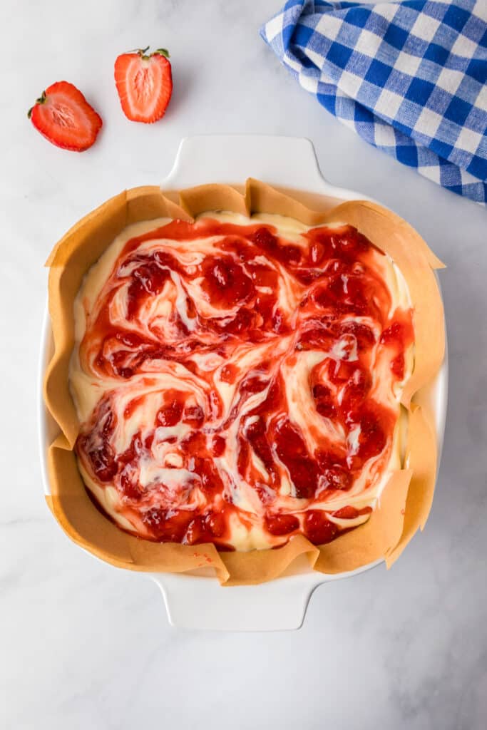 A baking dish with raw strawberry cheesecake bars swirled with with the strawberry mixture on a counter.