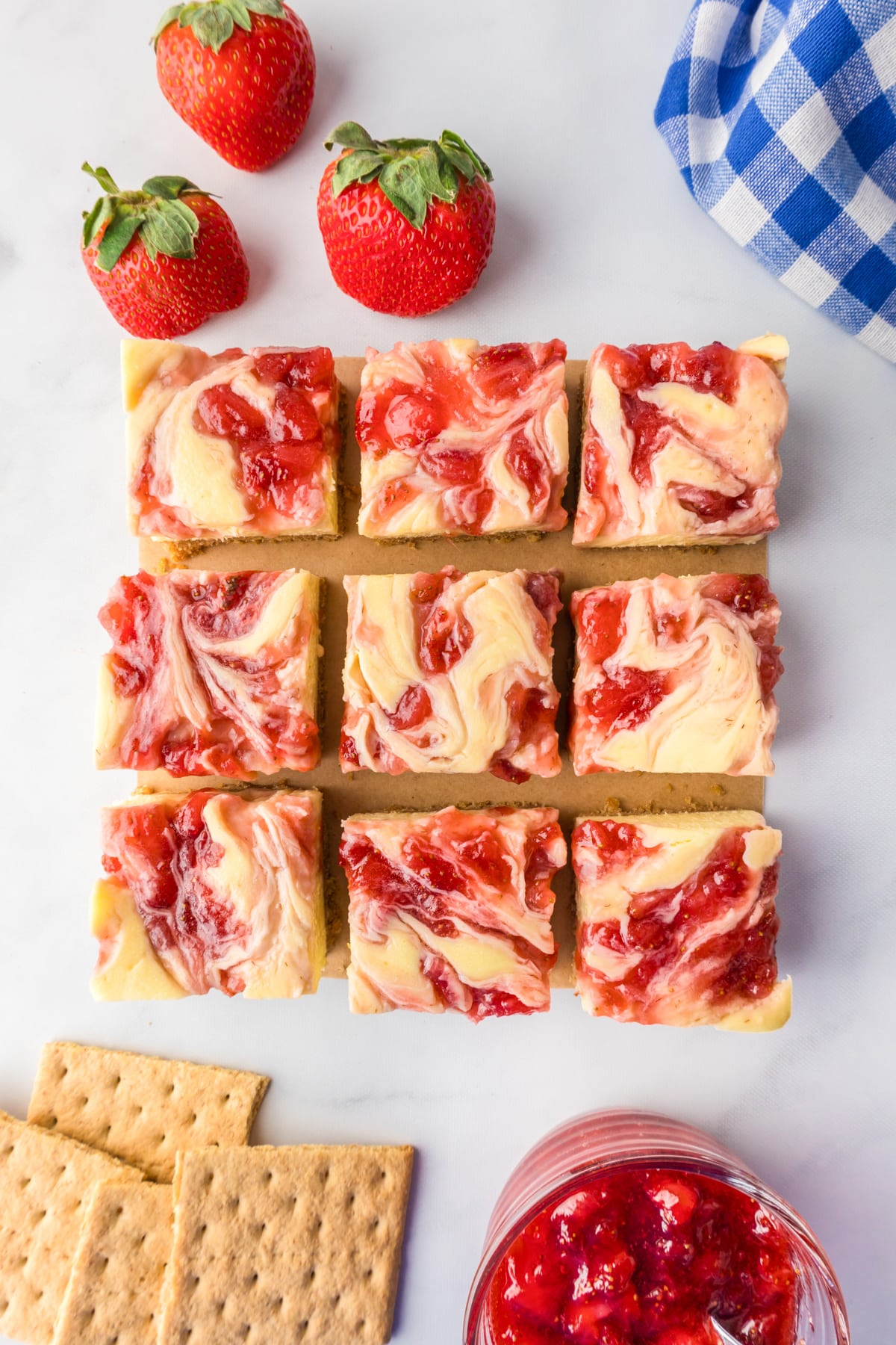 Nine strawberry swirl cheesecake bars arranged on a piece of parchment paper, with fresh strawberries, graham crackers, nearby.