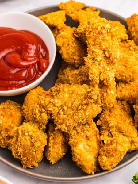 A plate of crispy breaded cornflake chicken nuggets being served with a bowl of ketchup for dipping.