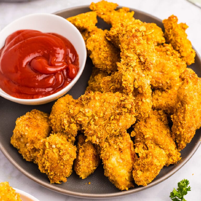 A plate of crispy breaded cornflake chicken nuggets being served with a bowl of ketchup for dipping.