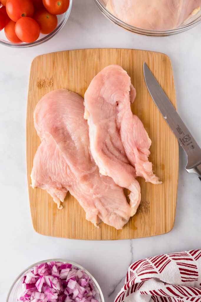 Two raw chicken breasts on a wooden cutting sliced into two cutlets next to a knife for greek chicken wraps.