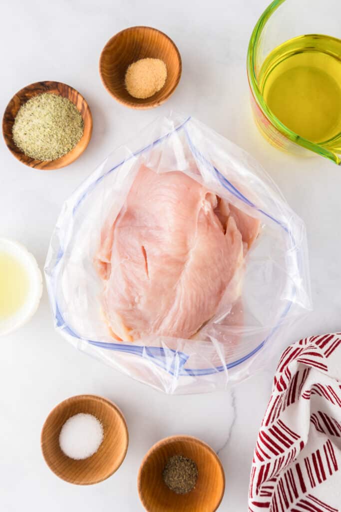 Raw chicken breast in a plastic bag surrounded by small bowls of seasoning and oil for a marinade for greek chicken.