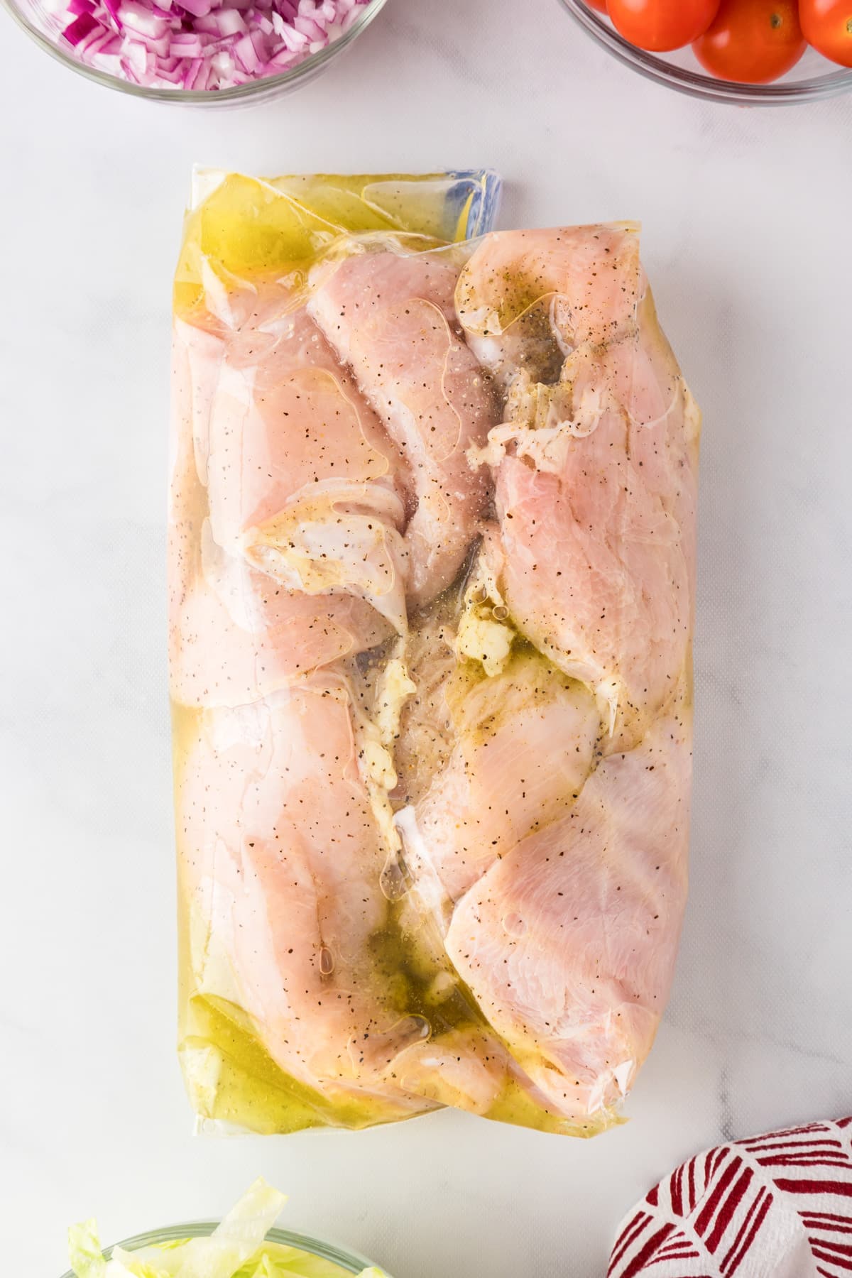 Raw chicken breasts marinating in a sealed plastic bag in a seasoned Greek marinade.
