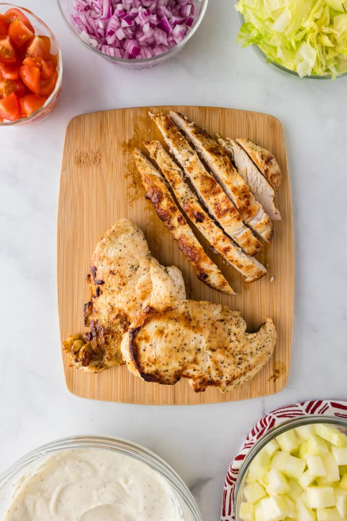 Sliced cooked greek chicken breast on a cutting board with more wrap ingredients and the greek yogurt sauce in bowls nearby.