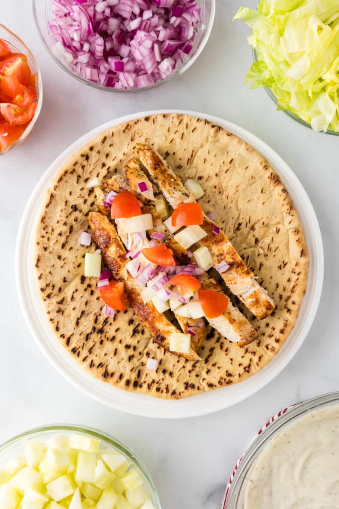 A pita bread topped with grilled Greek chicken strips, chopped tomatoes, onions, and cucumbers surrounded by more topping ingredients and the greek yogurt sauce in bowls.