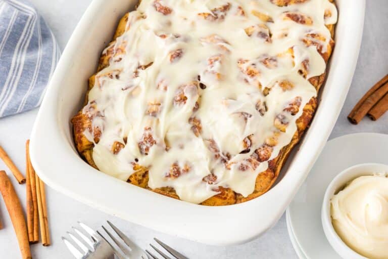 A baking dish filled with cinnamon rolls topped with cream cheese icing, next to a bowl of extra icing, cinnamon sticks, and forks on a white surface.