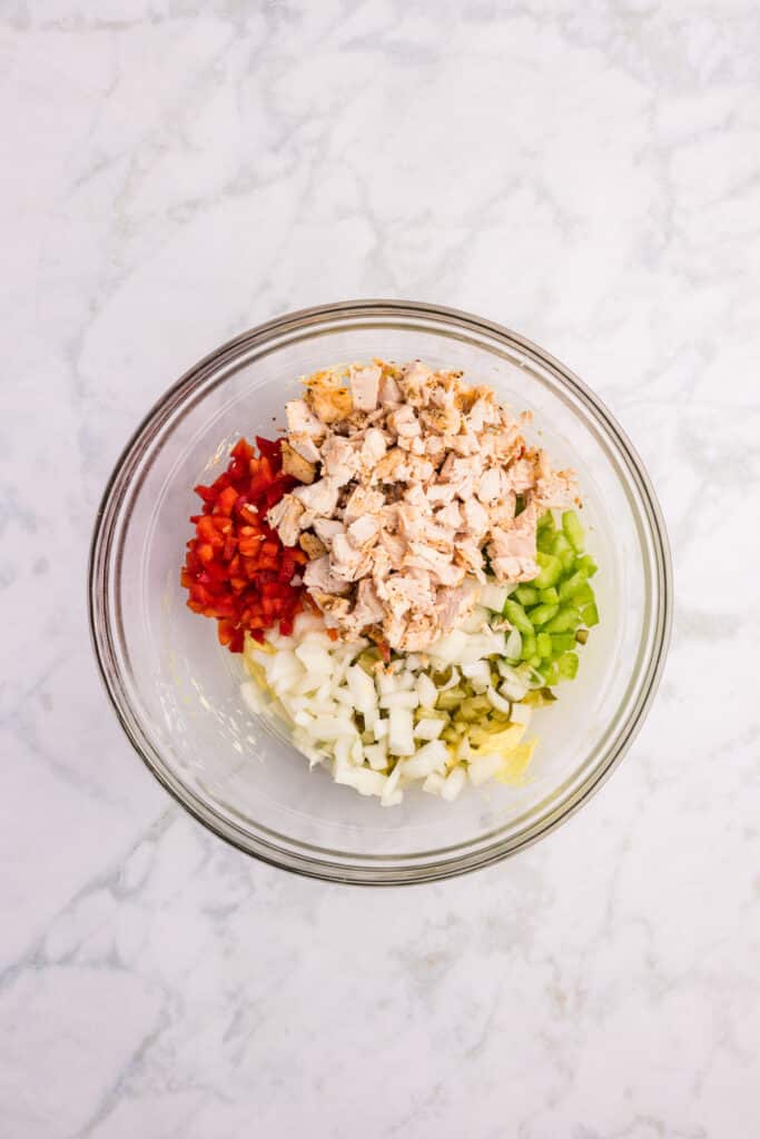 A glass bowl filled with chopped cooked chicken, diced red bell pepper, chopped celery, chopped pickles and diced onion for chicken macaroni pasta salad.