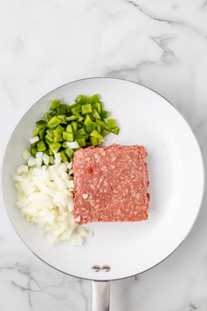 A white skillet contains raw ground beef, diced green bell pepper, and chopped onion on a marble countertop.