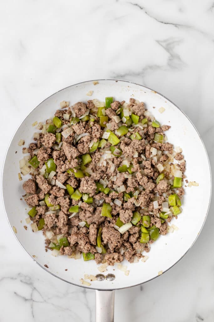 A skillet containing cooked ground beef, chopped green bell peppers, and diced onions on a marble countertop.