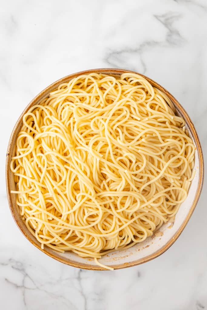 A bowl filled with plain cooked spaghetti noodles sits on a white marble surface.