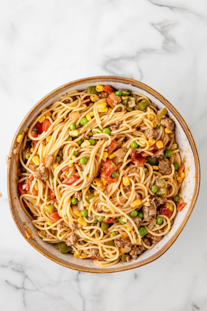 A bowl of spaghetti mixed with ground meat, corn, peas, diced tomatoes, and green bell peppers on a marble surface.
