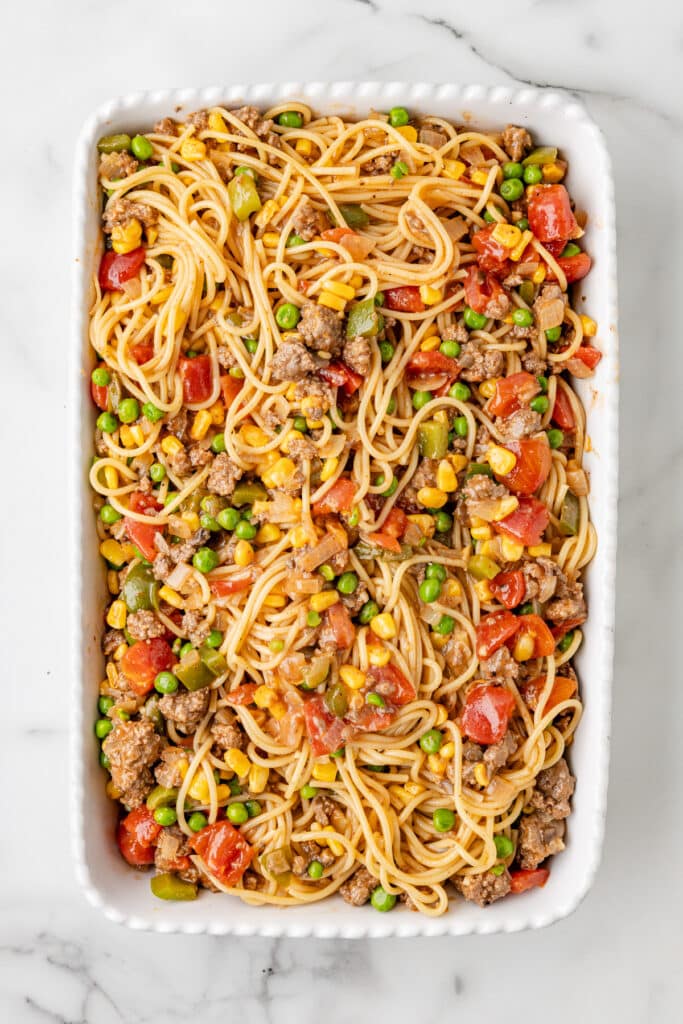 A rectangular white baking dish filled with spaghetti mixed with ground meat, peas, corn, diced tomatoes, and bell peppers.