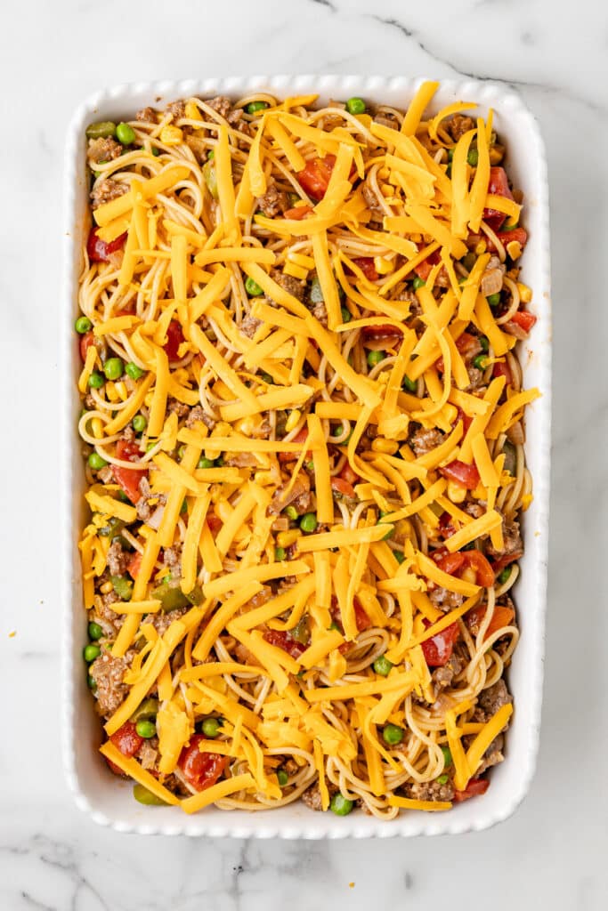 Rectangular baking dish filled with uncooked pasta, mixed vegetables, ground meat, and topped with shredded cheddar cheese on a marble surface.