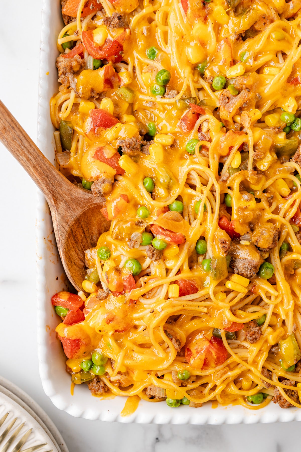Close up edge view of baked church super spaghetti in a casserole dish with a spoon scooping ground beef, peas, corn, pasta, diced tomatoes, and melted cheddar cheese.