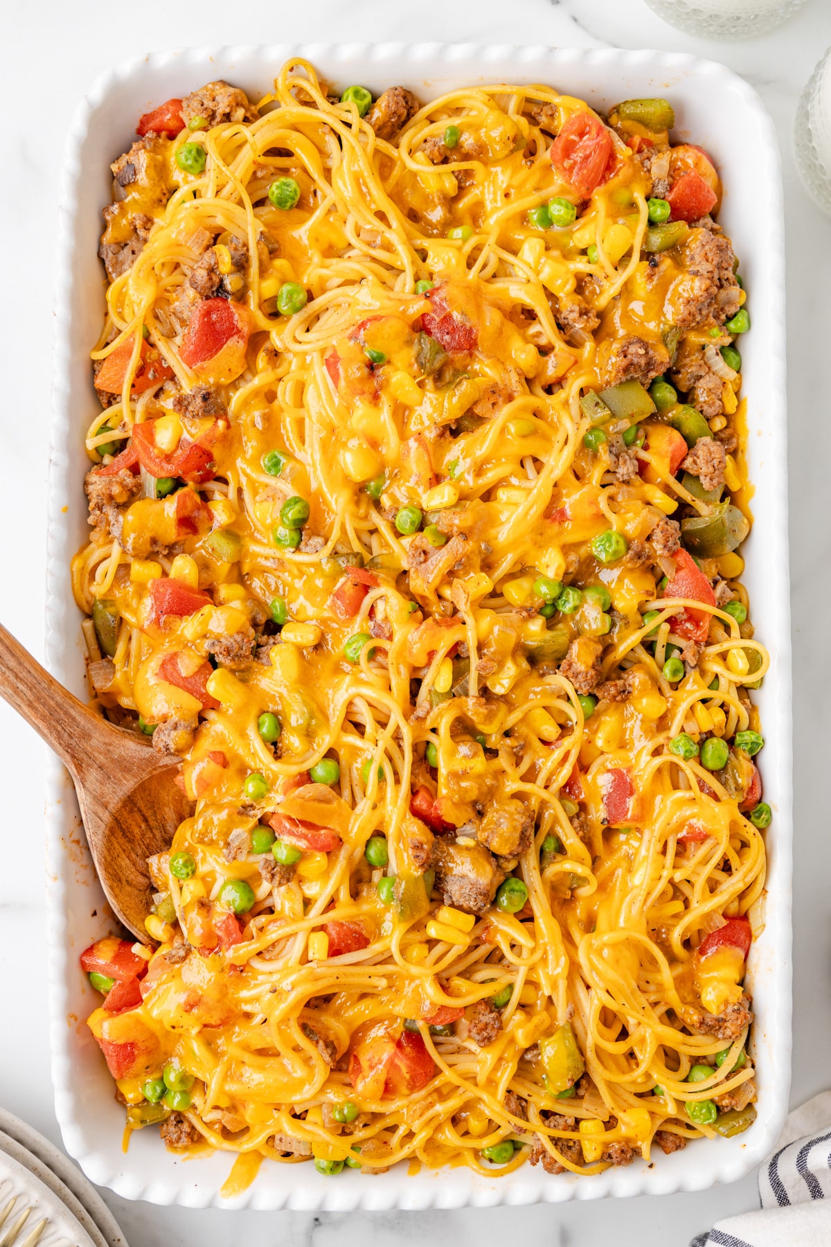 A rectangular baking dish filled with cheesy baked church supper spaghetti with a wooden spoon starting to scoop.
