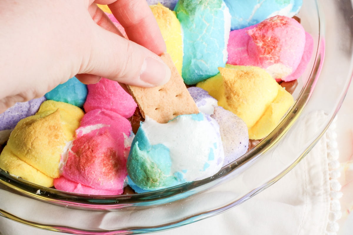A hand dips a piece of graham cracker into a colorful, swirled Peeps marshmallow dessert dip in a glass dish.