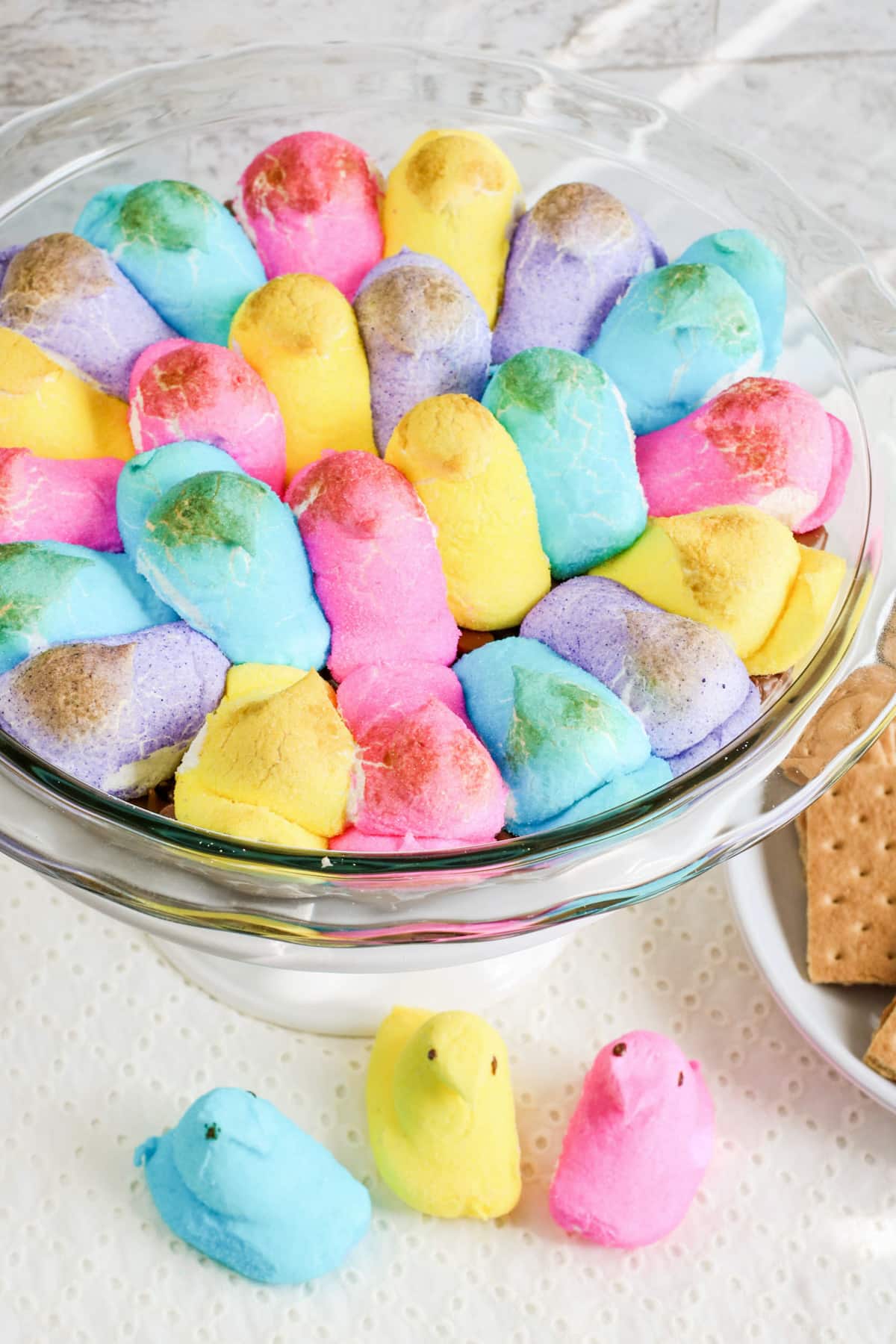 A glass pie pan full of Peep's smores dip with a few peeps chick marshmallows and graham crackers nearby.